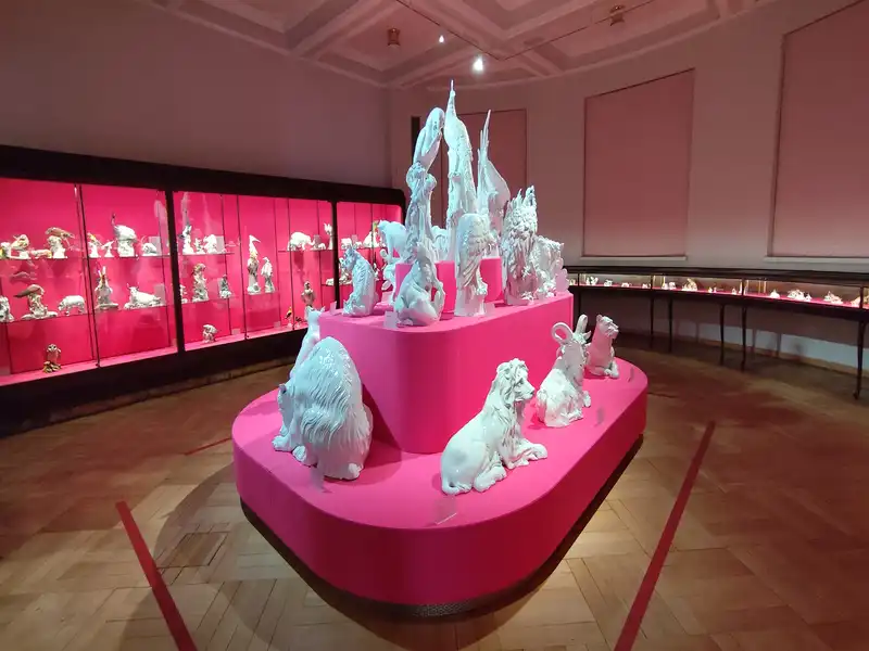 A Meisseni Porcelán Múzeum egyik terme Belső nézeti fotó a Meisseni Porcelán Múzeum egyik modern kiállítóterméről. A terem közepén egy nagyméretű, többszintes, lekerekített formájú, élénk magenta (neonrózsaszín) színű bemutatóállvány áll. Az állványon különböző méretű, rendkívül részletgazdag, tiszta fehér mázas porcelán állatfigurák láthatók, köztük egy fekvő oroszlán, egy bakkecske, egy jegesmedve és különféle madarak (például gémek és ragadozó madarak) a felső szinteken. A bal oldali fal mentén sötét keretes, szintén élénk magenta hátterű, kivilágított üvegvitrinek sorakoznak, amelyekben számtalan kisebb porcelánfigura kapott helyet. A jobb oldali falnál alacsonyabb, vízszintes üvegtárlók futnak végig. A terem padlója klasszikus halszálka mintás fa parketta, a mennyezetről pedig meleg fényű spotlámpák világítják meg a kiállítást, miközben az előtérben piros kordonvonalak jelzik a látogatók számára a biztonságos távolságot.