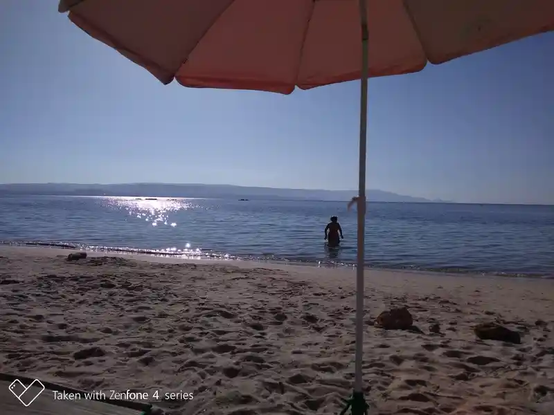 A beach scene on the Spiaggia del Lazzaretto beach in Alghero, photographed from under an open peach-colored parasol. In the foreground, the white pole of the parasol and the shaded part of the sandy beach with a few small rocks can be seen. In the background, the calm, blue sea stretches out, on which the sunlight shines brightly on the surface of the water. A lone human figure stands with his back turned in the shallow water, while in the distance, under a clear, blue sky, the silhouette of a mountainous coastline is outlined on the horizon.