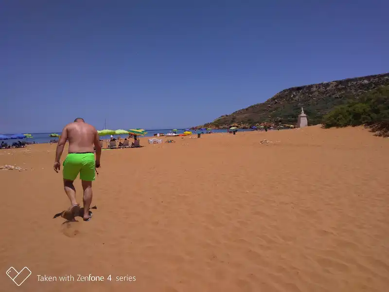 Csodálatos vörös homokos strand Gozo szigetén a Ramla Bay Napsütötte életkép a gozói Ramla Bay híres, jellegzetes vöröses-narancssárga homokos strandjáról. A kép bal előterében egy félmeztelen, neon zöld fürdőnadrágot viselő férfi sétál háttal a kamerának a széles, finomhomokos parton. A jobb oldalon egy zöldellő növényzettel és cserjékkel borított domboldal magasodik, amelynek szélén a jellegzetes fehér Mária-szobor emlékműve áll. A távolabbi partvonalon színes – kék, sárga és zöld – napernyők, napozóágyak és fürdőzők csoportjai láthatók a tengerpart mentén. A háttérben a nyugodt, kék tenger húzódik a teljesen tiszta, felhőtlen azúrkék égbolt alatt.