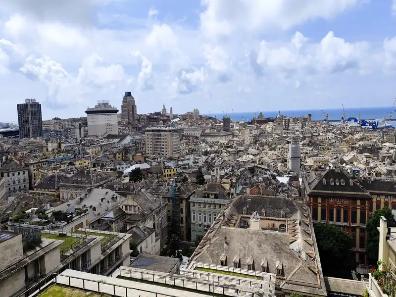 Genovai panoráma a Belvedere Castelletto kilátópontról Magaslati panorámakép Genova sűrűn beépített történelmi belvárosáról, a Belvedere Castelletto kilátópontról fotózva. Az előtérben régi bérházak és paloták változatos tetőszerkezetei láthatók szürke palatetővel, tetőablakokkal és néhány lapostetős, füvesített terasszal. A középtérben a történelmi negyed szorosan egymás mellé épült, világos homlokzatú házai és néhány modern, magasabb épület magasodik ki a városképből. A jobb oldalon egy díszes, vöröses homlokzatú, klasszikus palotaépület húzódik meg. A háttérben felsejlik a Földközi-tenger kék víztükre, ahol kikötői daruk és egy kék-fehér színű nagy hajó körvonalai látszanak. Az egész jelenet felett világoskék égbolt húzódik, amelyet sűrű, fehér, gomolyos felhők borítanak.