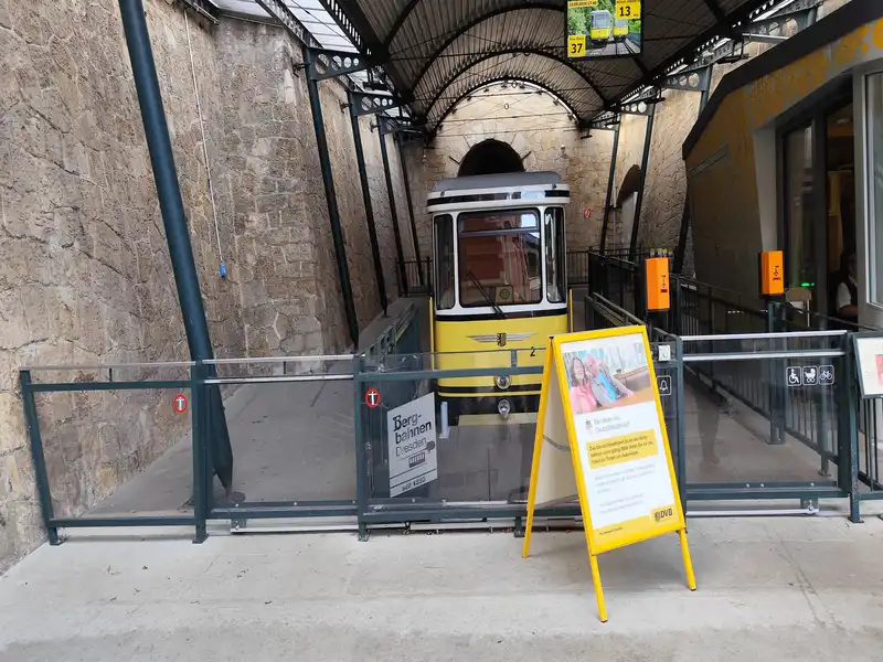 Fotó a drezdai sikló (Standseilbahn Dresden) egyik állomásáról. A kép közepén a sárga és fekete színű, lekerekített, retró kialakítású siklókocsi látható, amely éppen egy félköríves, sötét kőalagút bejárata előtt áll a síneken. Az állomást egy íves, sötét fémvázas tetőszerkezet fedi le, amelyről egy digitális utastájékoztató kijelző lóg. A bal oldalon magas, rusztikus kőfal húzódik, míg a jobb oldalon a peron bejárata látható két narancssárga jegykezelő oszloppal. Az előtérben, a biztonsági üvegkorlát előtt egy sárga keretes megállítóreklám áll német nyelvű plakáttal, rajta egy idős házaspár fotójával és a helyi közlekedési vállalat (DVB) logójával. Az üvegkorlát bal oldalán a „Bergbahnen Dresden seit 1895” feliratú információs tábla látható.