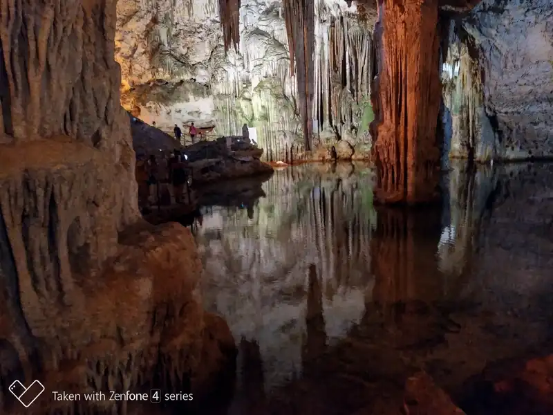 Alghero - Grotta di Nettuno, a belső sós vizű tó Egy lélegzetelállító, tágas cseppkőbarlang belsejéről készült felvétel. A kép középpontjában egy csendes, tiszta vizű földalatti tó látható, amely tökéletesen tükrözi a mennyezetről lógó és a talajból kiemelkedő monumentális cseppkőalakzatokat. A barlangot mesterséges, meleg sárga és zöldes-kék fények világítják meg, amelyek kiemelik a sziklák textúráját. A jobb oldalon egy masszív, narancssárga-barna cseppkőoszlop áll, amely mélyen a tó vizébe nyúlik. A bal oldalon, az előtérben egy vaskos, barázdált cseppkőoszlop takarja ki a kilátás egy részét. A háttérben, a tó túlpartján lévő megemelt járdán és lépcsőn apró emberalakok láthatók, amint megcsodálják a látványt. A mennyezetről több száz éles sztalaktit lóg lefelé, tükröződve a víz felszínén.