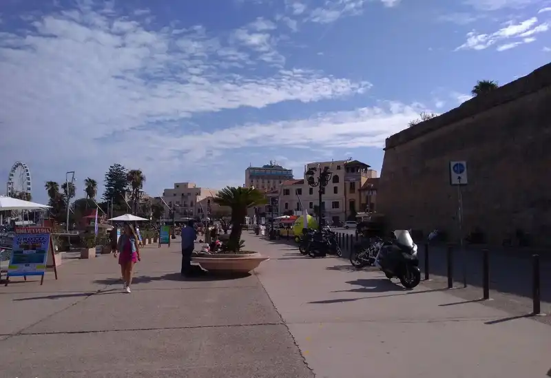 Alghero - Sétány a kikötőben a várfalakkal Széles, napfényes tengerparti sétány az olaszországi Alghero kikötőjében, a történelmi városfalak tövében. A kép jobb oldalán az óvárost védő masszív, magas kőfal magasodik, előtte parkoló motorkerékpárokkal és robogókkal. A térkövezett promenádon pálmafás dézsák és sétáló emberek láthatók, köztük a bal előtérben egy rózsaszín ruhás gyalogos nő, mellette kirándulásokat és barlangtúrákat hirdető kék-fehér reklámtáblával. A háttérben a város többszintes épületei, pálmafák és egy távoli óriáskerék sziluettje rajzolódik ki a kék, bárányfelhős égbolt alatt.